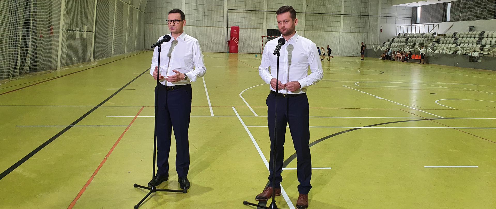 Premier Mateusz Morawiecki i Minister Kamil Bortniczuk ogłosili nowy pilotażowy Program budowy hal sportowych o lekkiej konstrukcji. Premier i Minister stoją przy mikrofonach w zadaszonej hali sportowej w Raszynie. W tle widoczne są biegające za piłką dzieci.