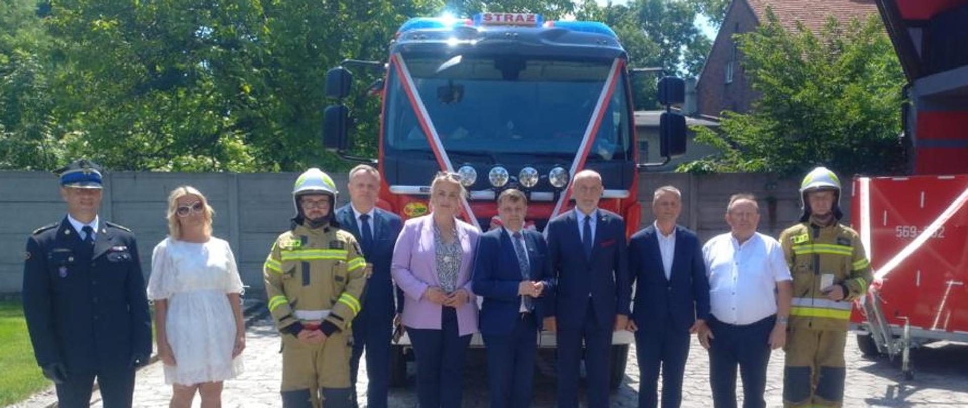 Grupa kobiet i mężczyzn stoi w rzędzie przed udekorowanym wozem strażackim