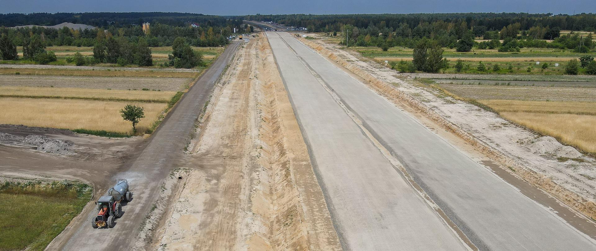 Na zdjęciu widać drogę w budowie oraz jadący traktor, a w oddali widać zieleń.
