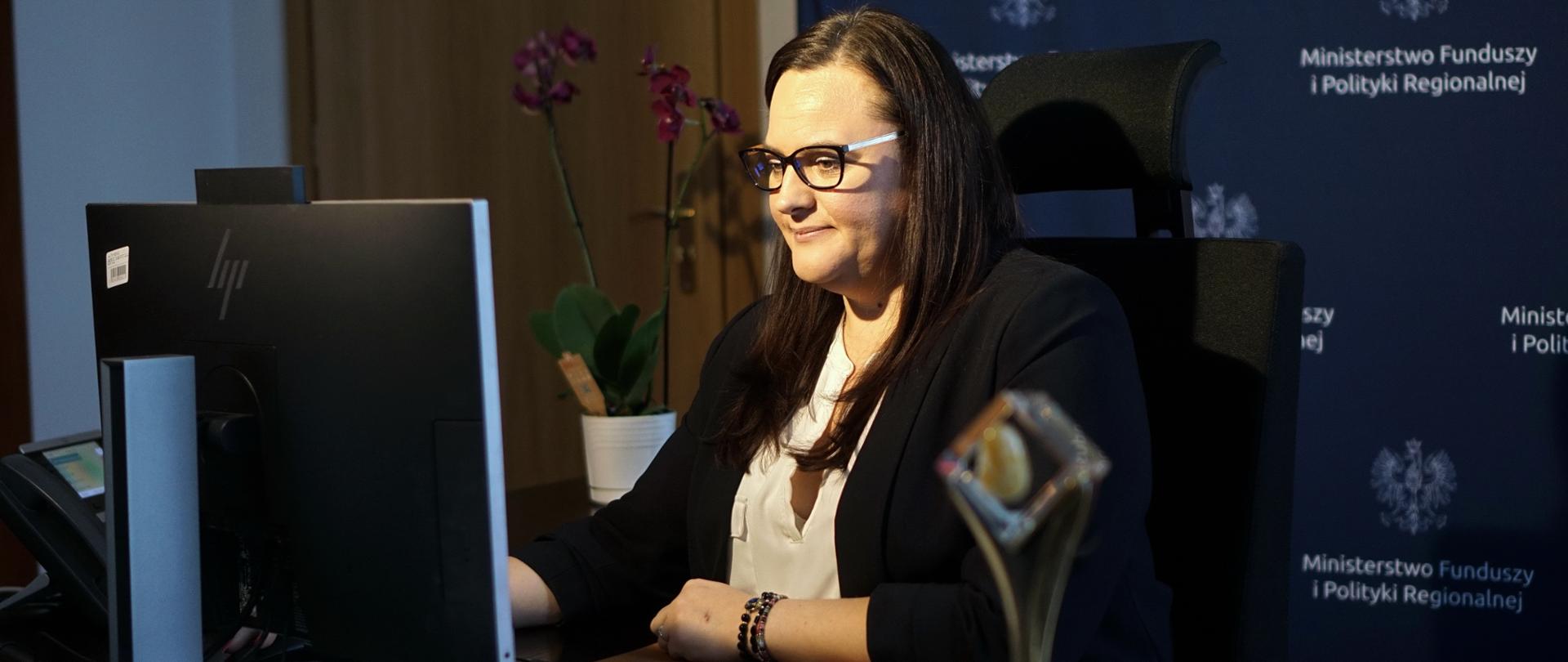Minister Małgorzata Jarosińska-Jedynak siedzi przy biurku przed monitorem komputera, w tle ścianka z logotypami Ministerstwa Funduszy i Polityki Regionalnej