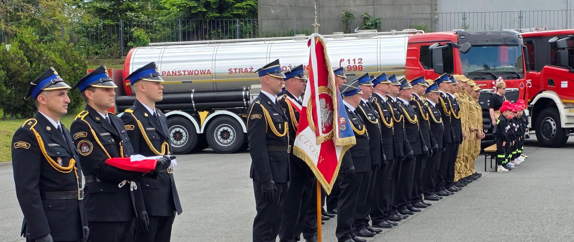 Uroczysty apel na placu komendy. W pierwszym planie z lewej strony stoi trzyosobowy poczet flagowy w ciemnych mundurach galowych, z których jeden trzyma złożoną flagę państwową. Za nimi stoi poczet sztandarowy ze sztandarem Komendy Powiatowej PSP w Głubczycach, a dalej w rzędzie wyrównany pododdział strażaków w mundurach wyjściowych oraz ubraniach koszarowych piaskowego koloru. Na samym końcu szyku stoi grupa małych dzieci w miniaturowych strojach strażackich i czerwonych czapkach. W tle widoczny jest nowy, czerwony ciągnik siodłowy ze srebrną naczepą-cysterną z napisem "Państwowa Straż Pożarna" oraz fragment innego wozu strażackiego.