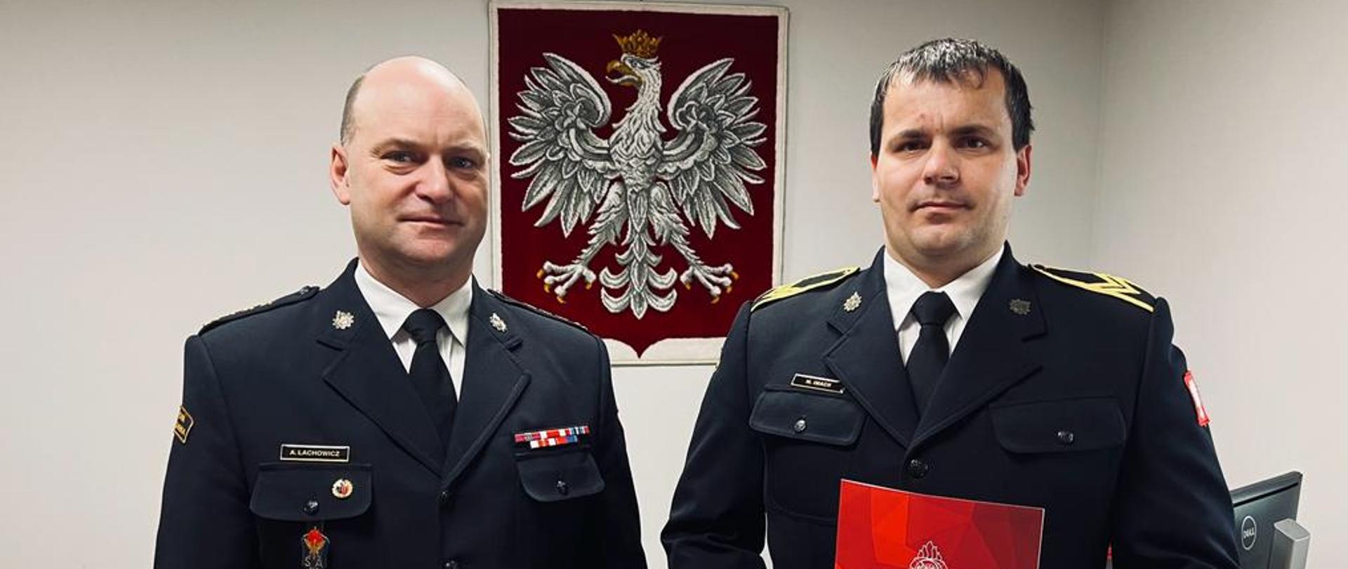 Dwóch strażaków stoi w mundurach wyjściowych. Za plecami flaga rzeczypospolitej polskiej