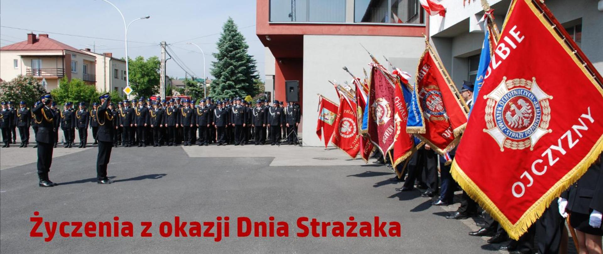 Na zdjęciu widzimy napis Życzenia z okazji Dnia Strażaka, w tle widzimy strazaków oraz sztandary.