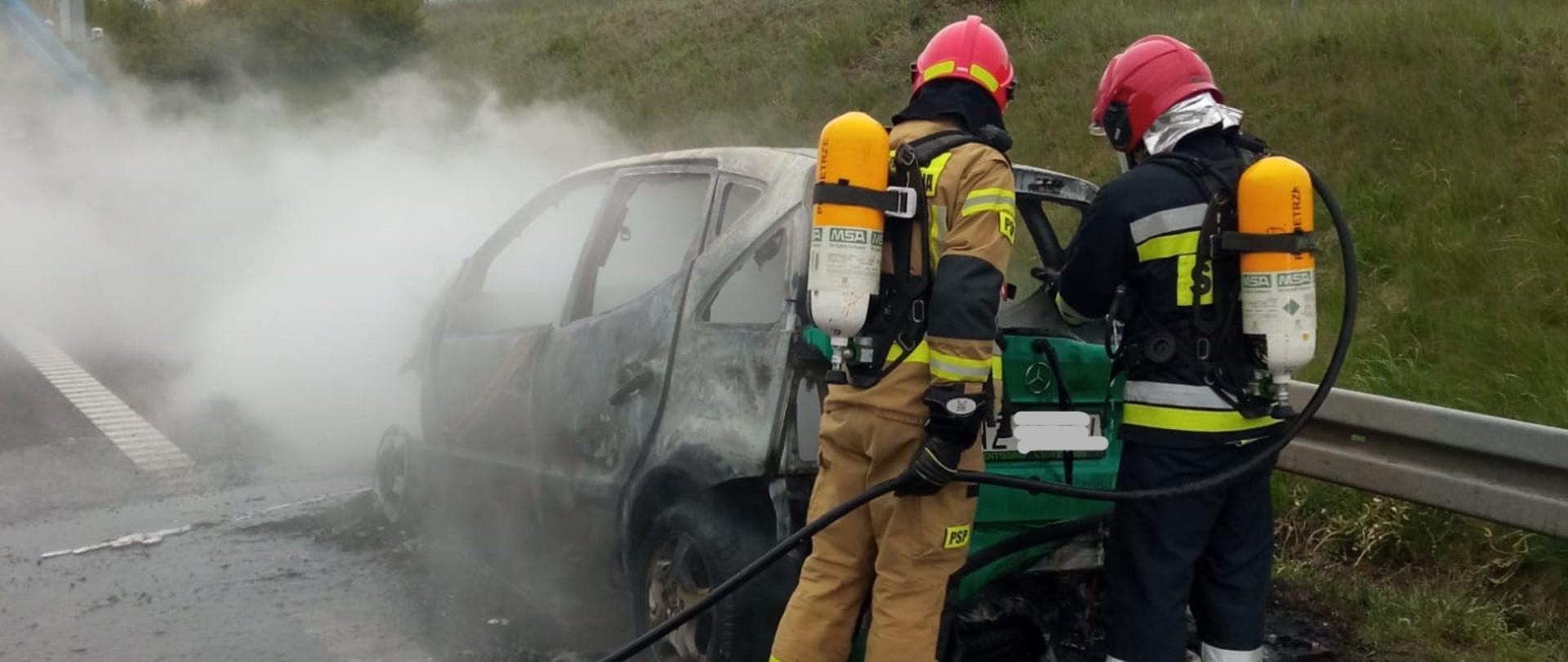 Zdjęcie przedstawia dwóch strażaków w aparatach ochrony układu oddechowego dogaszających spalony samochód stojący na pasie awaryjnym drogi S7.