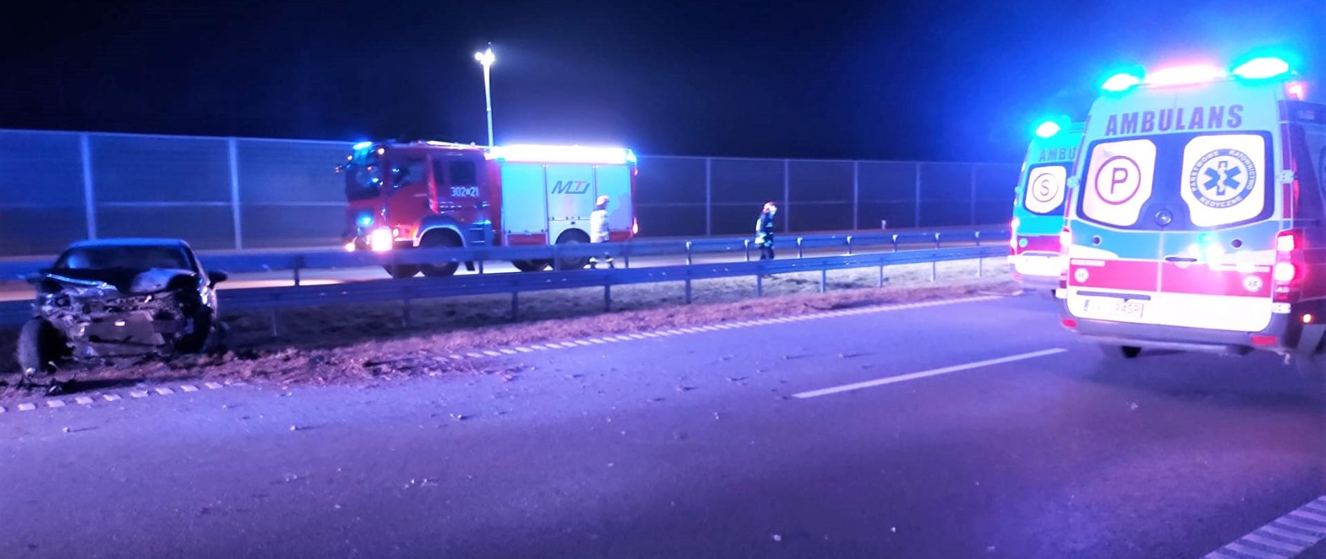 Na zdjęciu przedstawiono miejsce działań strażaków. Po lewej stojący przodem na pasie zieleni znajduje się samochód osobowy po wypadku. W centralnej części zdjęcia widać samochód pożarniczy a najbardziej po prawej stoją dwa ambulanse Ratownictwa Medycznego.