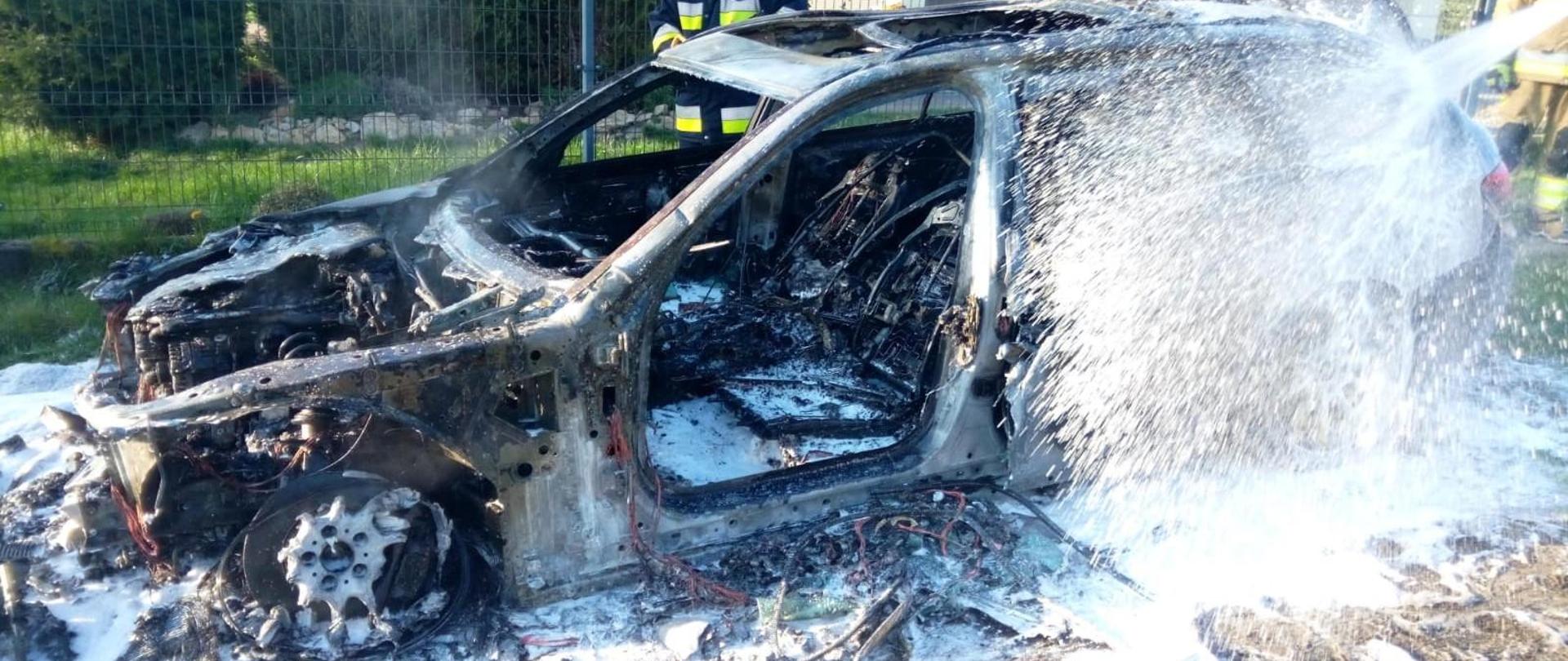 Zdjęcie przedstawia spalony samochód osobowy w trakcie dogaszania pożaru. Jest obficie pokryty pianą, która wciąż jest podawana z prawej strony.