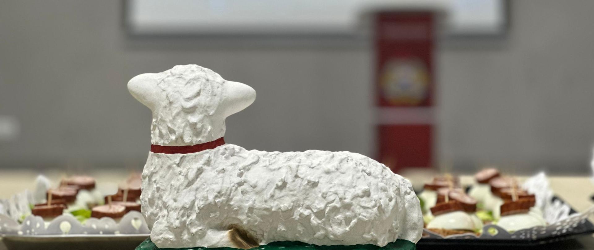 Na zdjęciu znajduje się baranek wielkanocny który leży na stole - figurka. Tło jest zamazane.