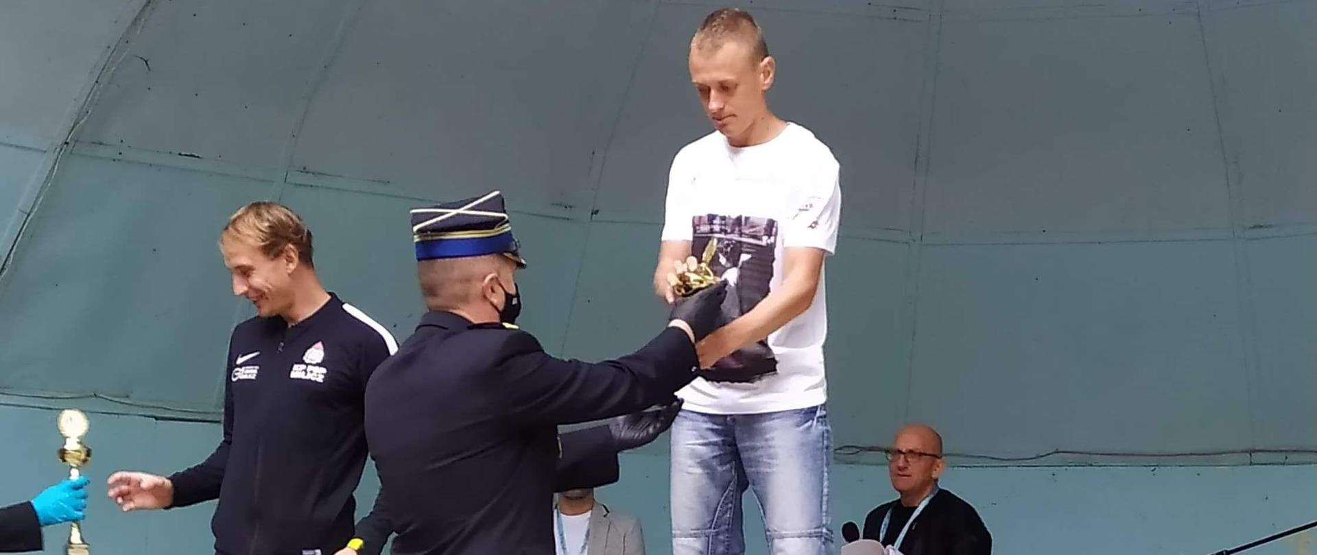 Zdjęcie przedstawia Dekoracja po biegu Zaślubin. Na zdjęciu widzimy stojących na podyższeniach - podestach uczestników biegu oraz wręczającego statuetki komendanta.