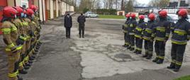 Zdjęcie przedstawia strażaków stojących w szeregu ubranych w piaskowe mundury oraz czerwone hełmy przed nimi stoją natomiast w szeregu strażacy w czarnych ubraniach specjalnych i hełmach, za nimi znajdują się czerwone bramy garażowe.