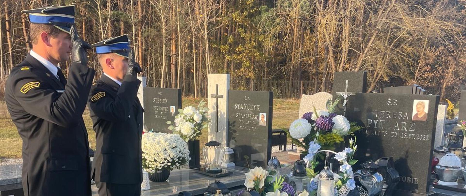 Strażacy w umundurowaniu wyjściowym stoją frontem do grobu na cmentarzu i oddają honor.