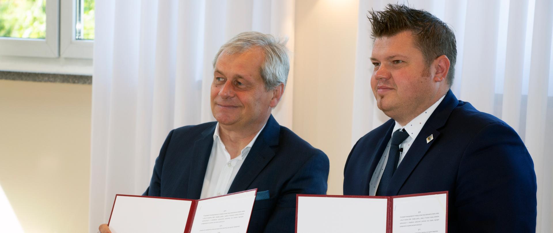 Na zdjęciu po lewej stronie kadru stoi wojewoda opolski. Mężczyzna w granatowym garniturze i białej koszuli, ma siwe włosy. w Dłoniach trzyma teczkę. Teczka jest otwarta, widać jej zawartość. To podpisana umowa. Obok wojewody stoi młodszy mężczyzna, ma ciemne, dłuższe włosy. Pozuje z wojewodą do zdjęcia. W jego dłoniach również widać teczkę.