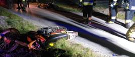 Zdjęcie przedstawia dwa motocykle crossowe leżące na poboczu drogi asfaltowej. Obok znajduje się kilku strażaków w umundurowaniu bojowym. Zdjęcie zrobione w porze nocnej.