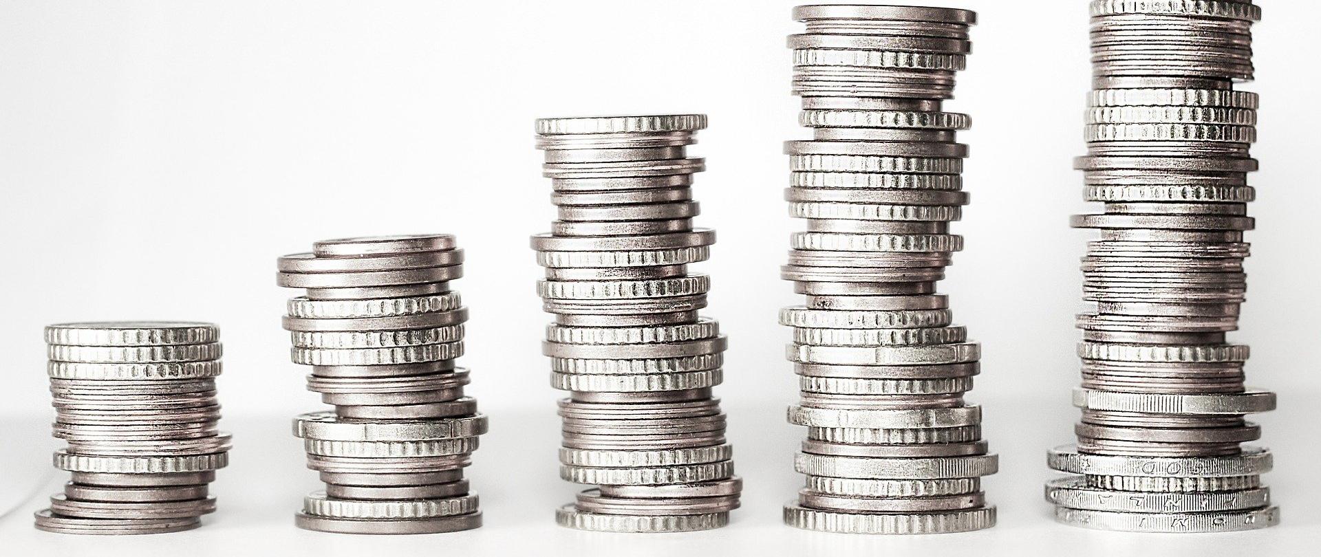 Na zdjęciu pięć "kupek" monet. Od lewej najmniejsza ich liczba, po prawej stronie najwyższa i największa. Monety ułożone na białym stole, w tle białe tło. 
