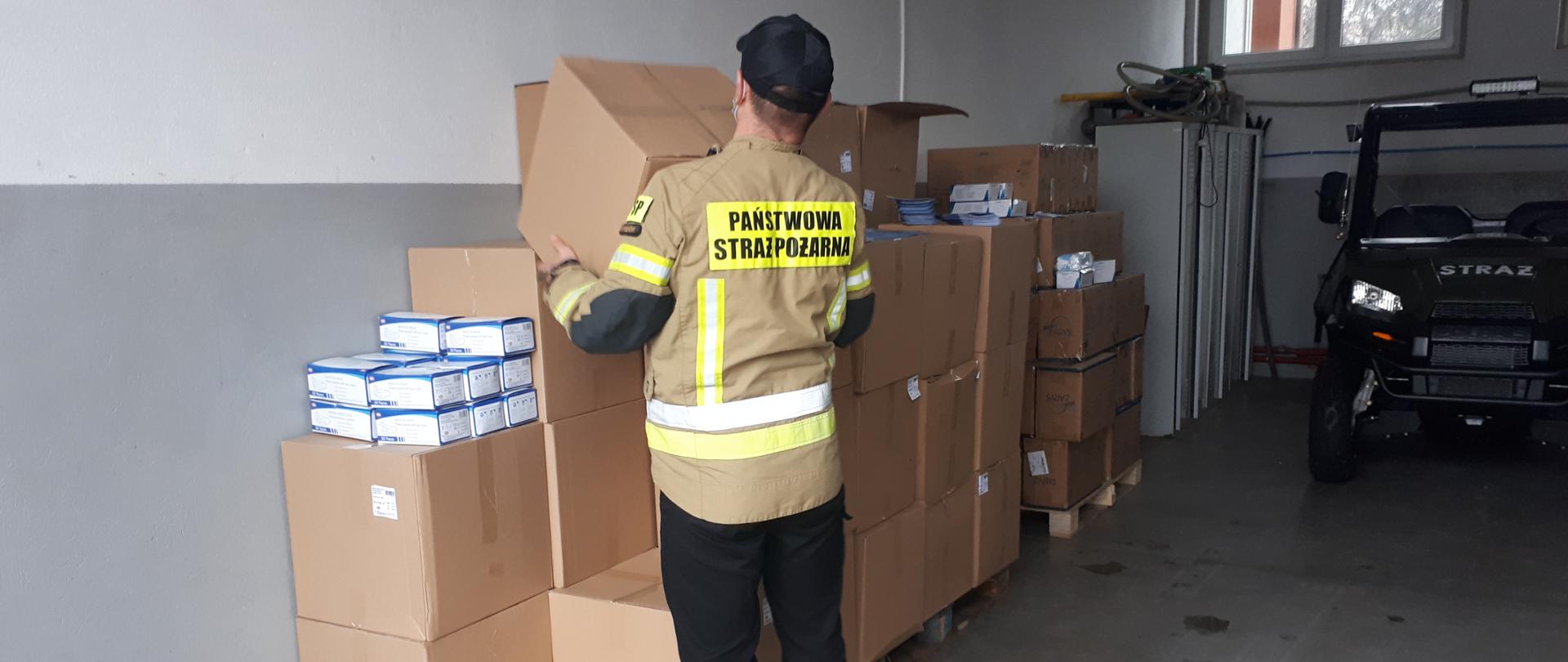 Zdjęcie przedstawia strażaka noszącego kartony z maseczkami ochronnymi