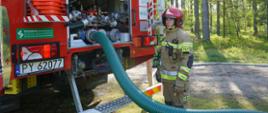 Na tle lasu stoi ciężki samochód pożarniczy. Za nim strażak, który sprawdza możliwość poboru wody ze zbiornika podziemnego. Do samochodu podłączona jest linia ssawna. Strażak ma na sobie czerwony hełm oraz ubranie specjalne.