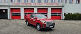 Czerwony strażacki pojazd typu pickup widziany od przodu z boku, stojący na utwardzonym placu przed szarym budynkiem czerwonymi bramami z napisem „Państwowa Straż Pożarna”