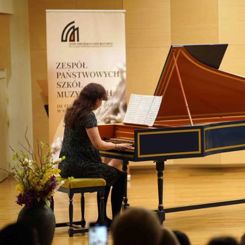 zdjęcie przedstawia uczennicę grającą na klawesynie na scenie sali koncertowej, w tle baner z logo szkoły