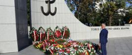 Na dworze w ostrym słońcu pod wielkim pomnikiem z symbolem Polskiego Państwa Podziemnego i napisem Tobie Ojczyzno - Armia Krajowa stoi mężczyzna w garniturze, pod pomnikiem leży dużo kwiatów z biało-czerwonymi wstążkami.