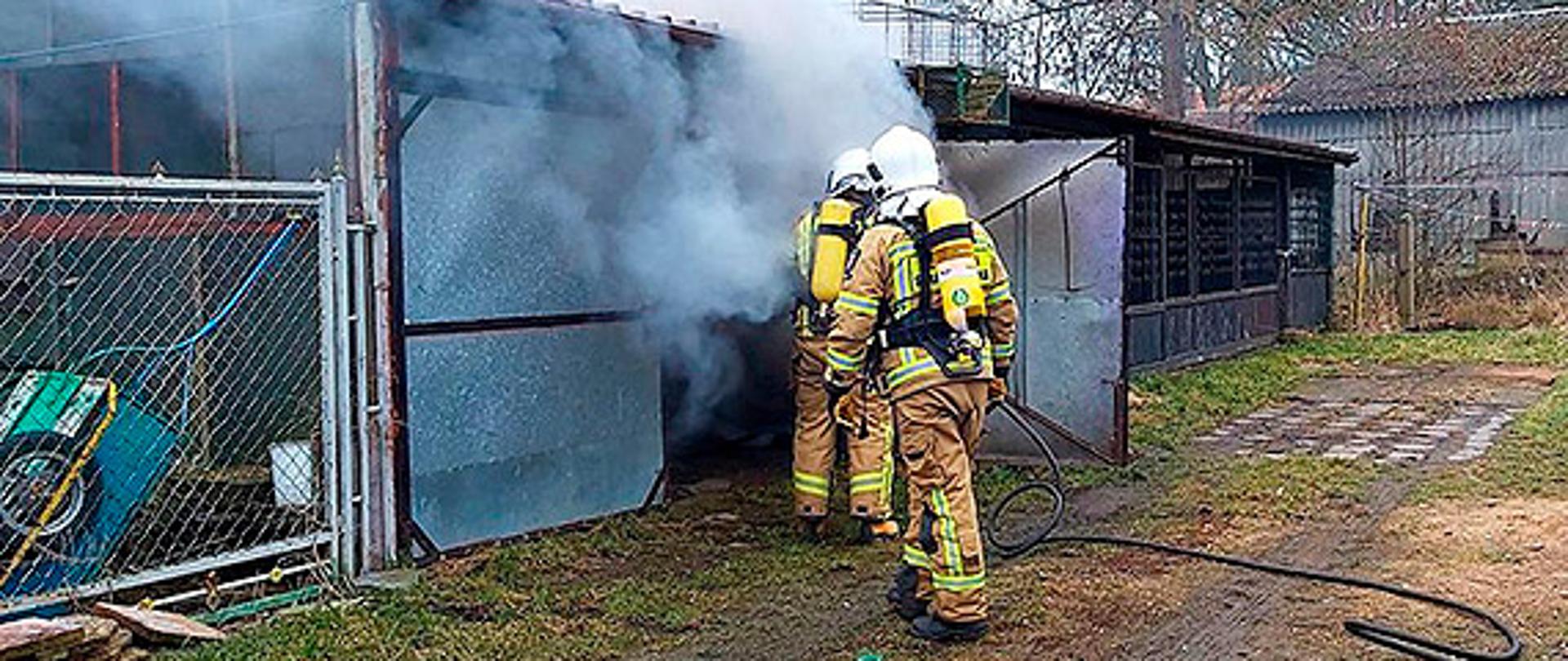 Zdjęcie przedstawia dwóch strażaków ubranych w aparaty powietrzne którzy wchodzą z linią gaśniczą do blaszanego garażu z którego wydobywa się dym.