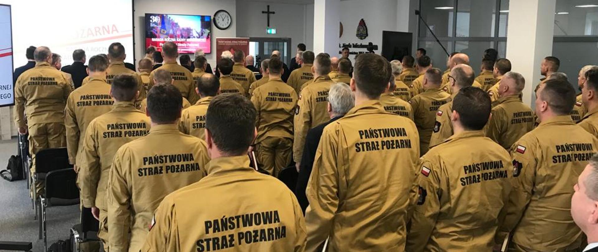 Widok z tyłu sali na której siedzi grupa strażaków. W oddali widoczny ekran z wyświetlana prezentacją