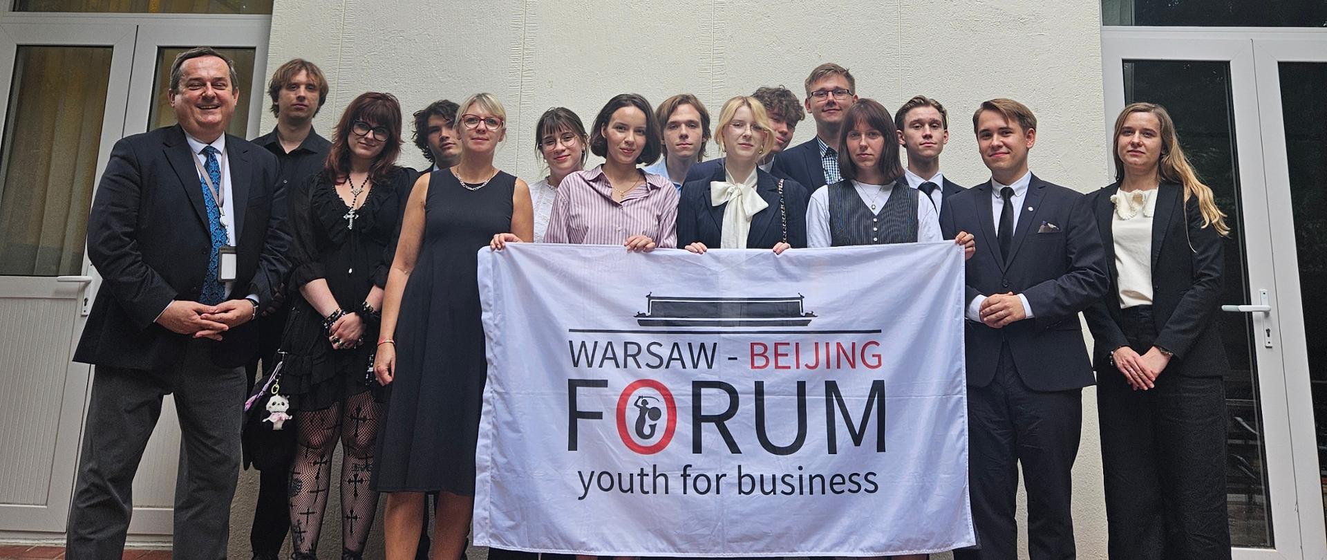 Grupa osób trzymająca flagę Warsaw-Beijing Forum stoi przed budynkiem konsulatu, pod flagami Polski i UE