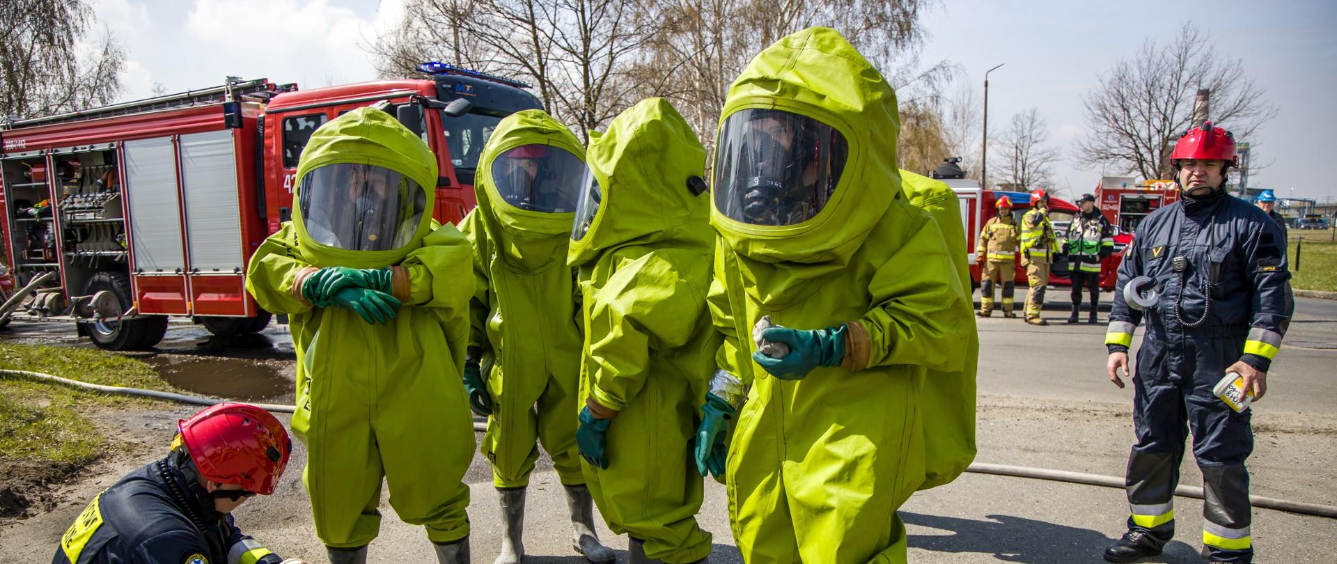 Strażacy PSP zabezpieczeni w ciężkich ubraniach gazoszczelnych czekają na wejście do strefy niebezpiecznej (podczas ćwiczeń). Ubranie chemiczne koloru zielonego. Pozostali strażacy przygotowują materiał do uszczelnienia wycieku na zbiorniku. W tle widoczni inni strażacy oraz samochody strażackie.