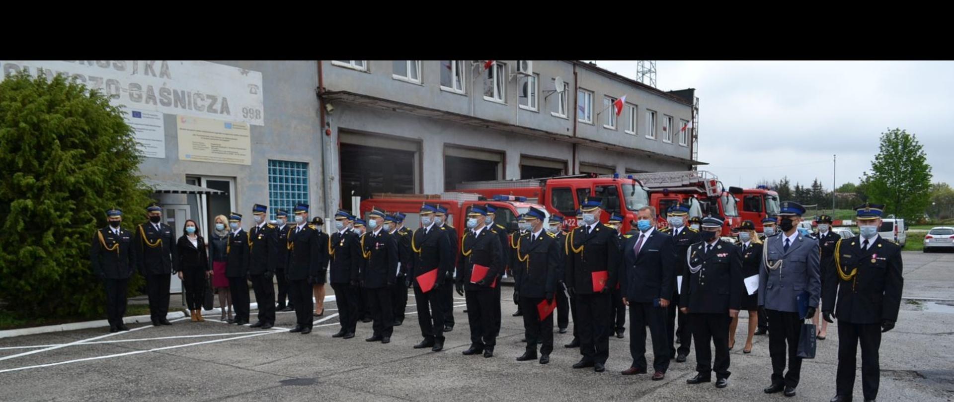Na planie widoczni są funkcjonariusze i funkcjonariuszki, którzy zostali nagrodzeni lub odznaczeni z okazji Dnia Strażaka 2021 z zaproszonymi gośćmi. W tle widoczny jest budynek Jednostki Ratowniczo – Gaśniczej w Żarach i samochody ratowniczo – gaśnicze przed garażami.