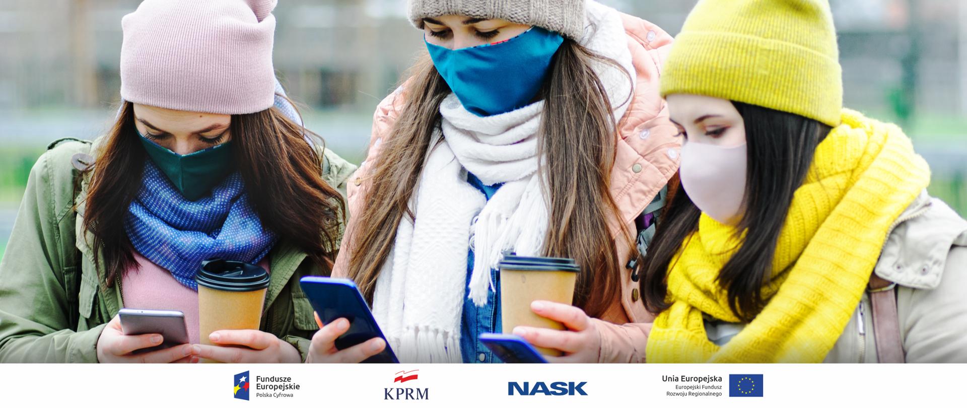 Trzy młode dziewczyny w jesiennych strojach i maseczkach - każda z nich trzyma w ręku smartfona i patrzy na jego ekran.
