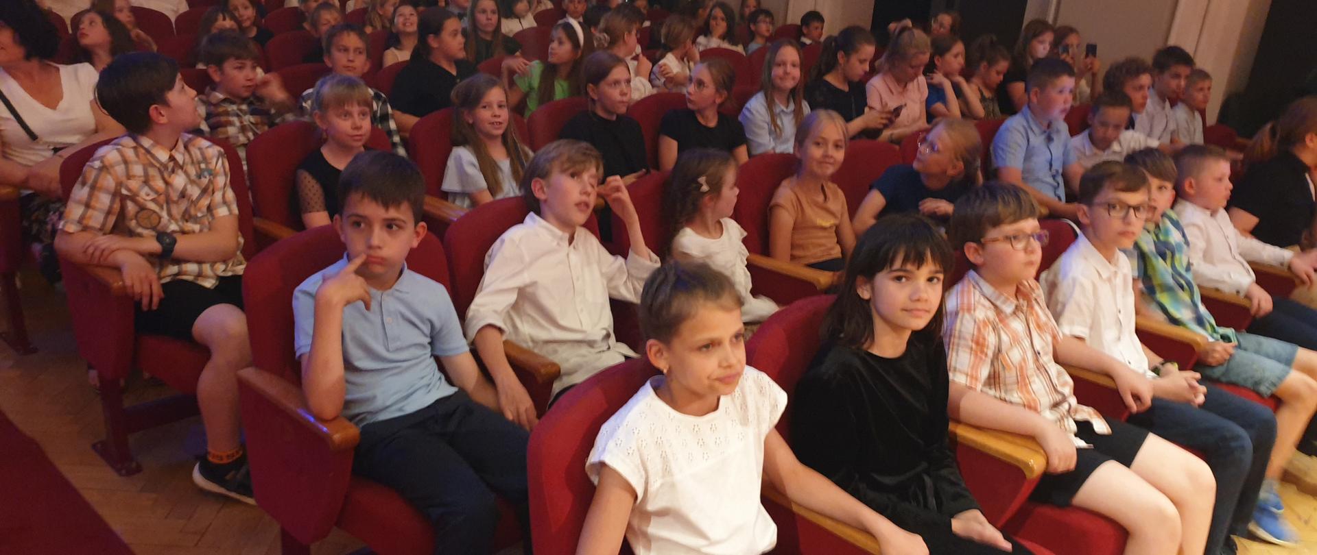 Widok na widownię z czerwonymi fotelami zapełnioną dziećmi.