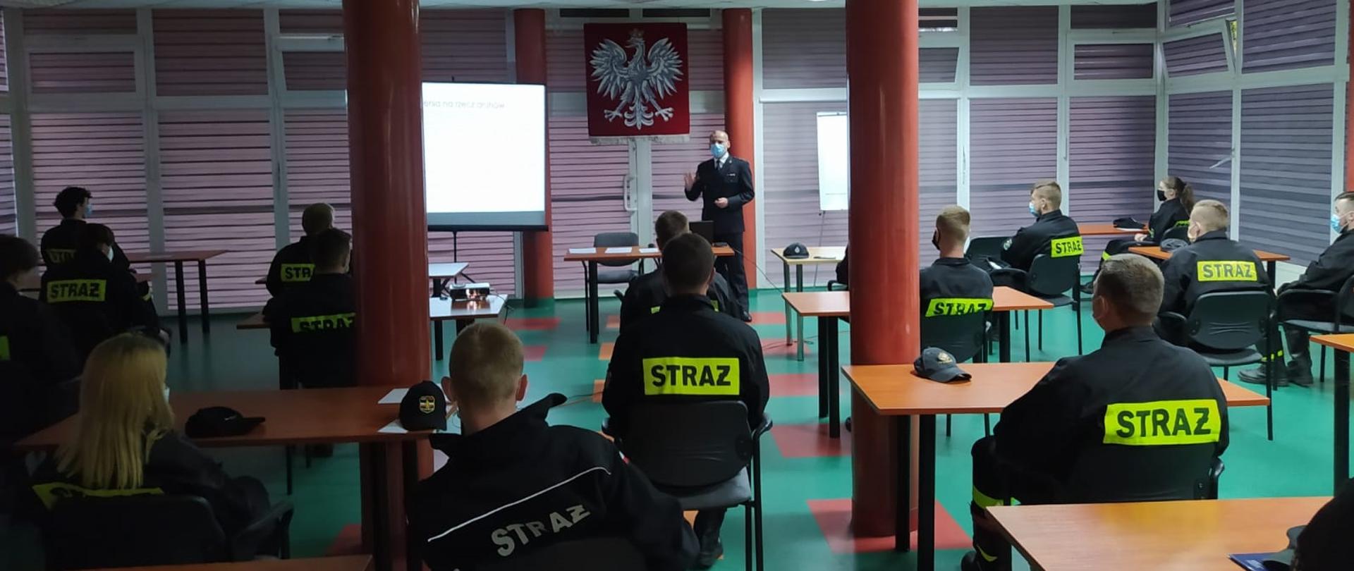 Na zdjęciu w centralnej części stoi starszy kapitan Damian Łojek w umundurowaniu wyjściowym. Po prawej jego stronie widoczne Godło Polski a następnie stronie ekran z wyświetloną prezentacją. Przed nim druhowie w umundurowaniu koszarowym siedzą na krzesłach w odstępach około 1,5 m. 