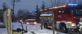 Pojazdy ratowniczo-gaśnicze ustawione przed budynkiem w którym doszło do pożaru kotłowni. Pojazdy maja włączone oświetlenie zewnętrze i niebieskie lampy błyskowe.