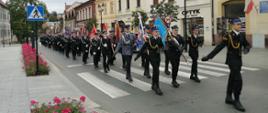 Zdjęcie przedstawia strażaków z Państwowej i Ochotniczej Straży Pożarnej w mundurach wyjściowych ze sztandarami, którzy maszerują w kolumnie ulicą Bochni. W tle znajdują się zabudowania miejskie