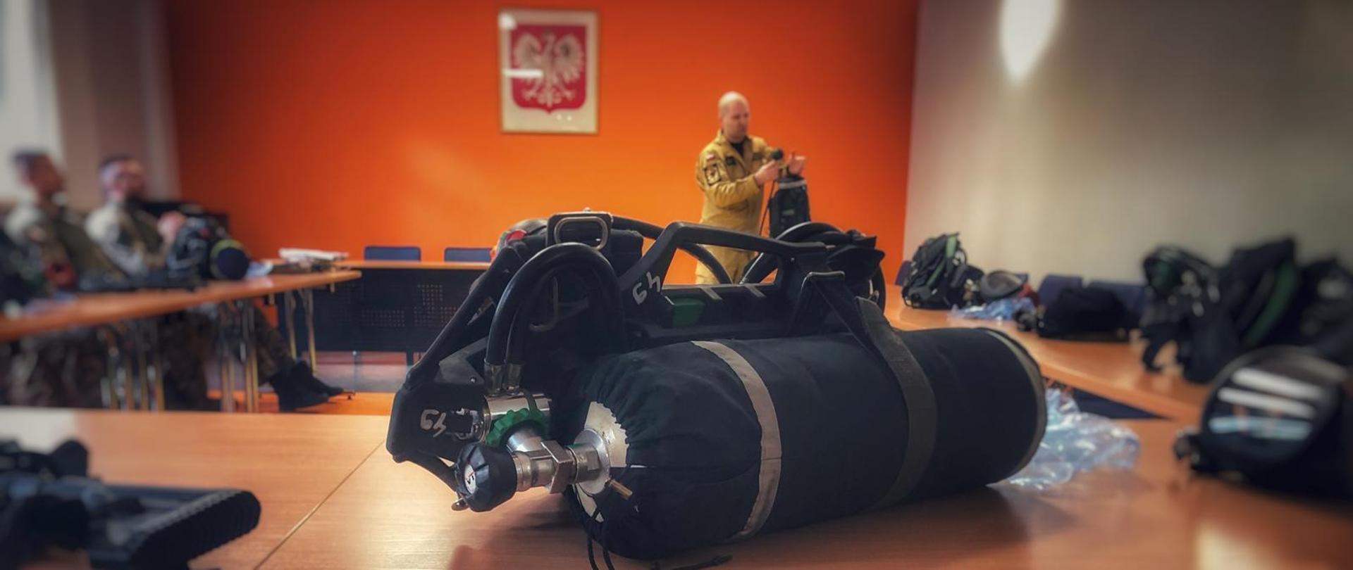 Na stole leży aparat dróg oddechowych w tle strażak demonstruje i omawia budowę sprzętu za stołami siedzą funkcjonariusze Policji.