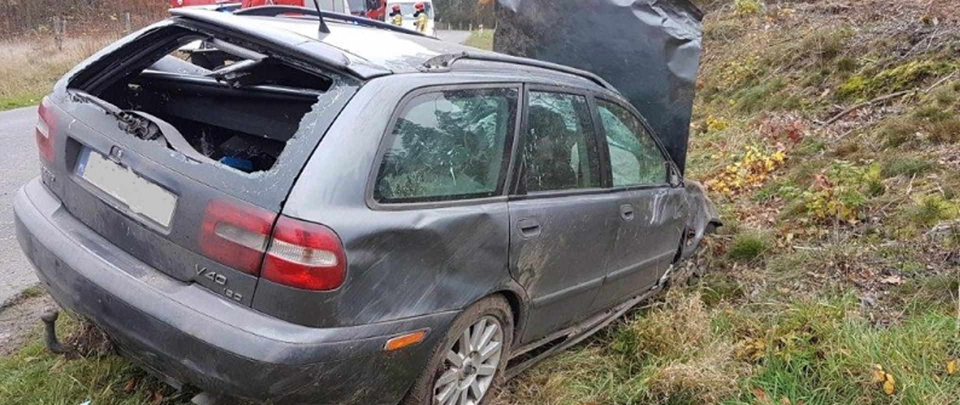 Zdjęcie przedstawia wnętrze tył samochodu osobowego Volvo koloru szarego stojący na kołach na poboczu drogi. Tylna szyba bagażnika wybita, bok samochodu od strony pasażera posiada liczne ślady otarcia lakieru. Maska silnika podniesiona do pionu. Za pojazdem widoczne dwa pojazdy strażackie i karetka pogotowia. Na tle karetki stoą dwaj strażacy w ubraniach specjalnych