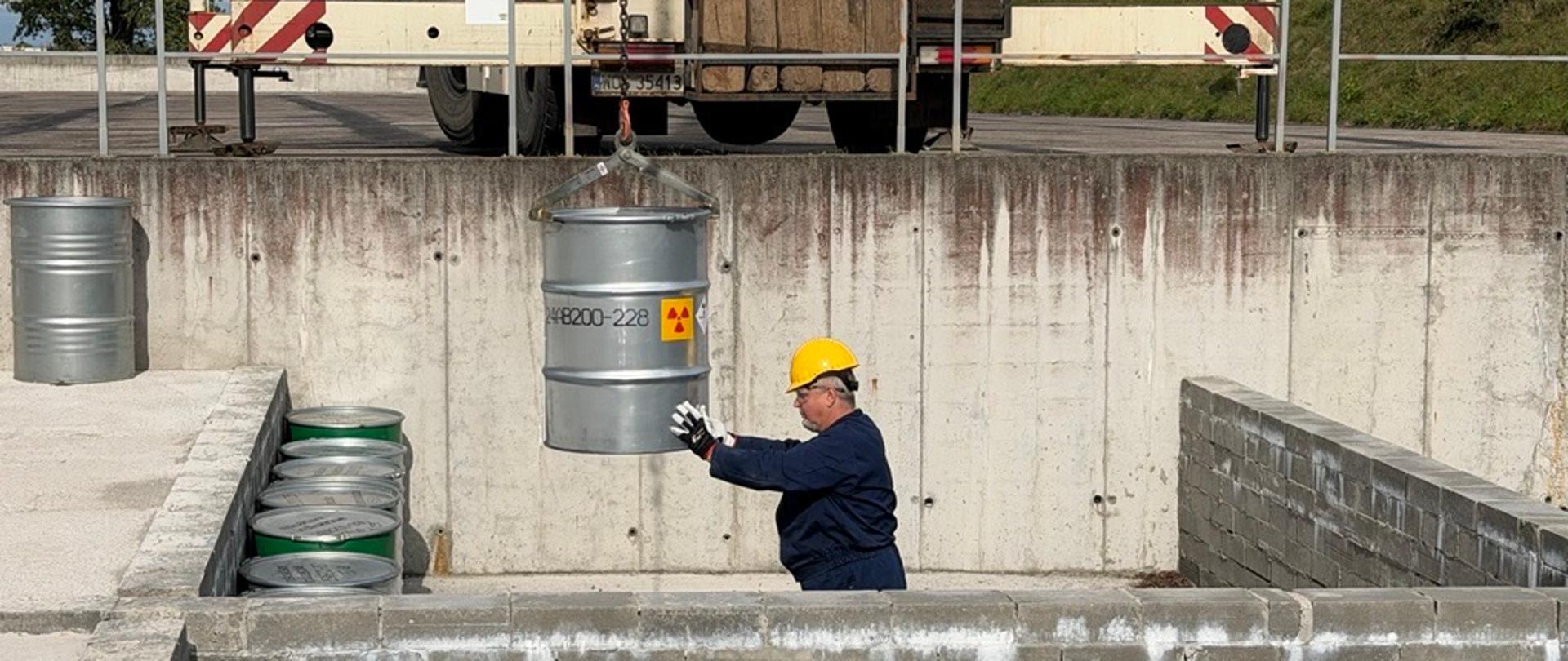 Pracownik ZUOP ustawia w sektorze stalowy stalowy bęben z odpadami promieniotwórczymi. Bęben opuszcza dźwig.