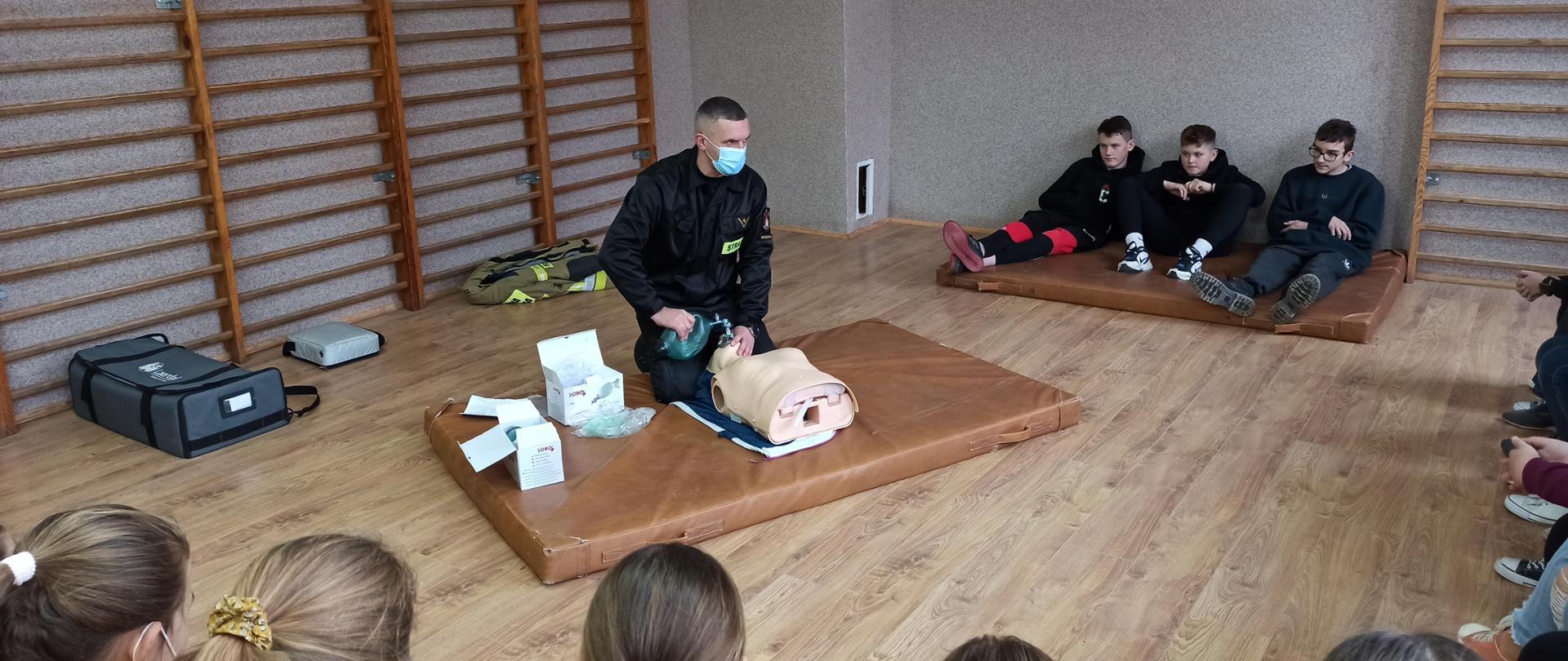 Mł. asp. Paweł Skarżyński podczas prezentacji technik ratowniczych. Dookoła dzieci oglądające prezentację.