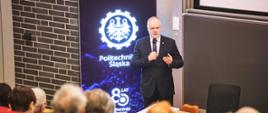 Zdjęcie od góry, wiceminister Gzik stoi na sali i mówi do trzymanego w ręku mikrofonu, za nim pionowy baner z napisem Politechnika Śląska na niebieskim tle.