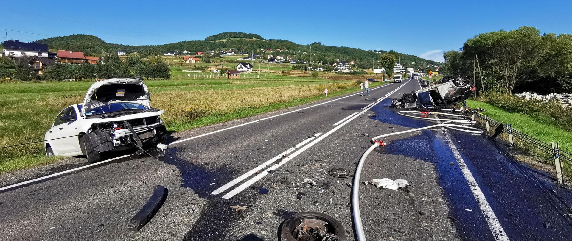 Samochody po zdarzeniu na drodze. Jeden po jednej stronie drogi na kolach a drugi na dachu. Urwane koło leży na drodze oraz rozwinięta linia gaśnicza.