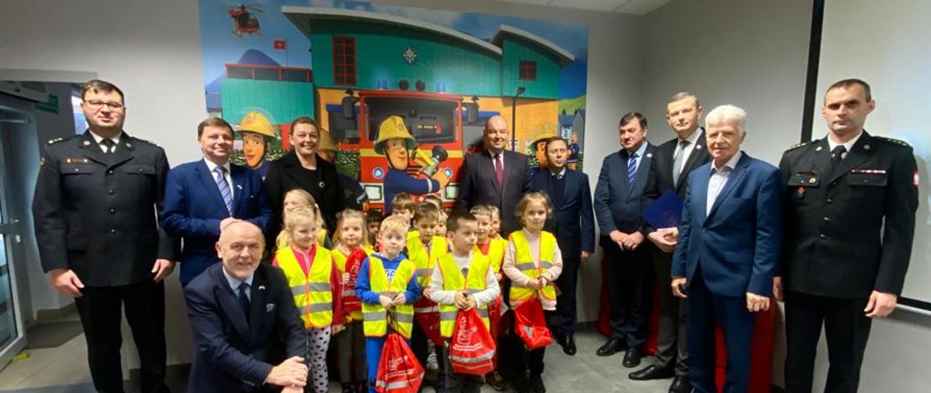 zdjęcie grupowe strażaków w ciemnych mundurachm osób cywilnych oraz dzieci w żółtych kamizelkach, w tle rysunek strażaka sama iczerwonego samochodu