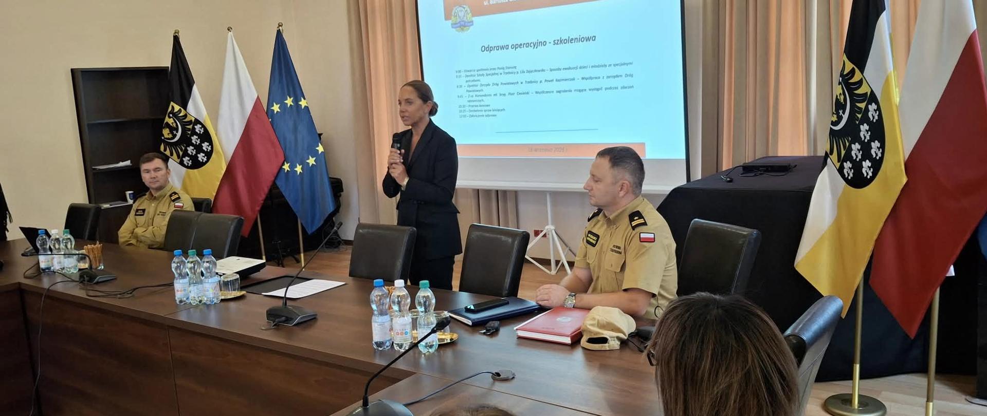 Podczas odprawy operacyjno-szkoleniowej Komendy Powiatowej Straży Pożarnej w Trzebnicy starosta powiatu, kobieta w czarnym żakiecie, przemawia do mikrofonu, stojąc przy długim stole między ubranymi w mundury komendantami, a na ścianie za nimi wisi duży ekran z polskimi flagami i emblematami.