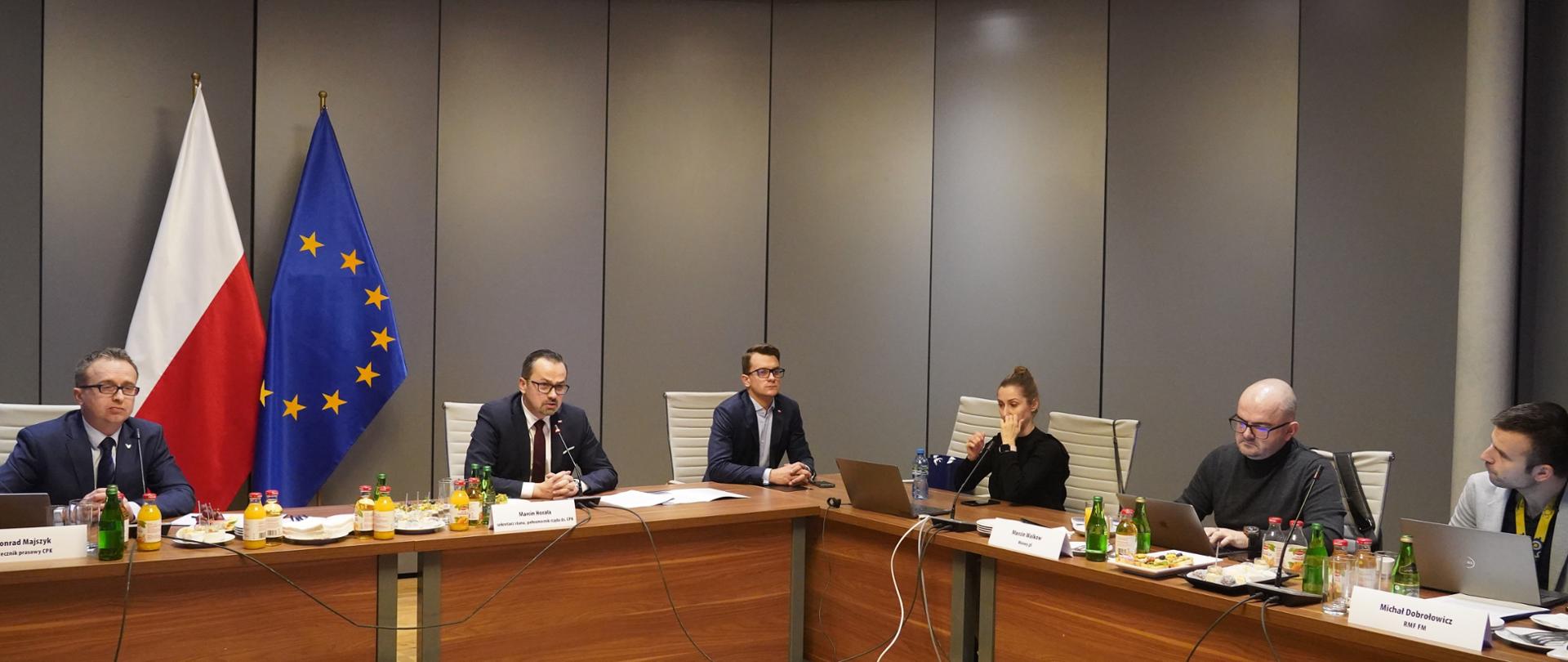 Sześć osób siedzi przy prostokątnym stole. Drugi od prawej siedzi wiceminister Marcin Horała. W tle za jego plecami stoją flagi Polski i Unii Europejskiej. 