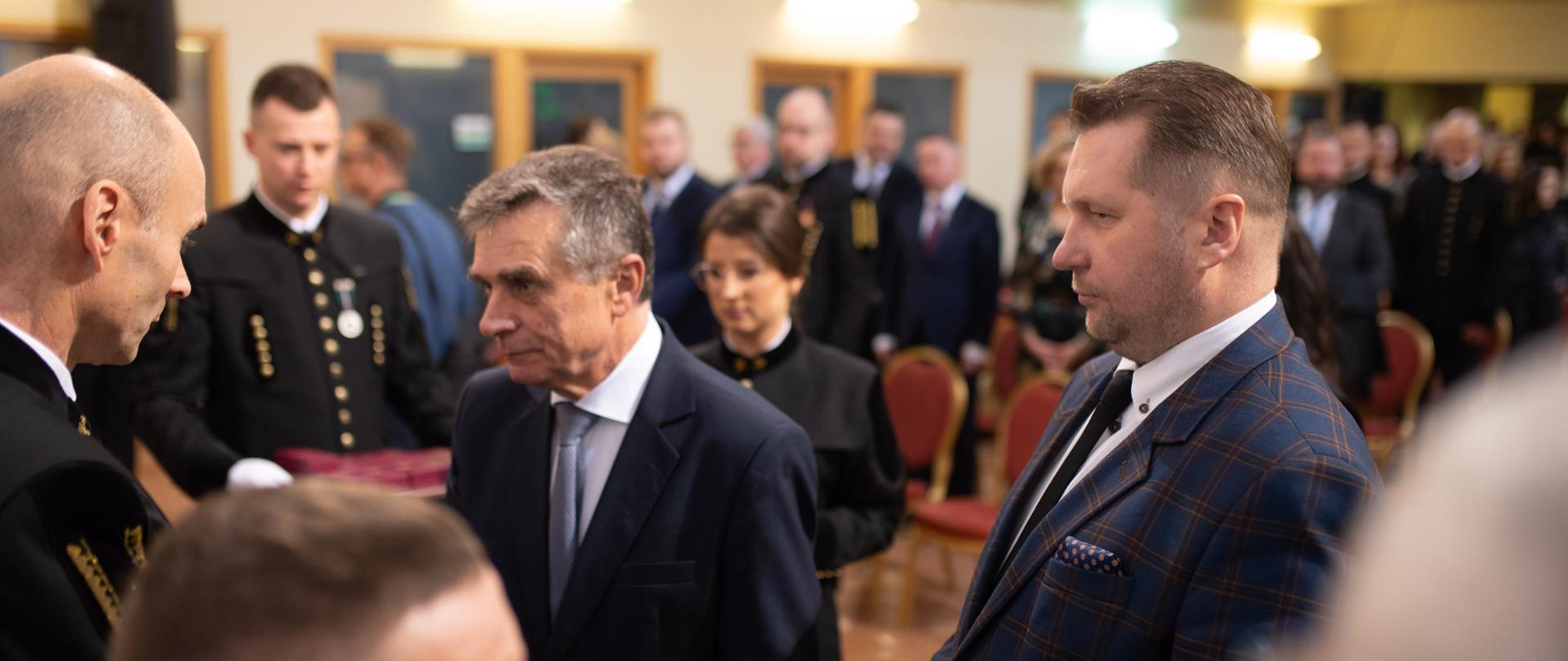 Minister Czarnek i stojący obok starszy mężczyzna w garniturze rozmawiają z ludźmi w czarnych galowych mundurach, minister trzyma w ręku otwartą małą książeczkę, dalej sala z żółtymi ścianami wypełniona ludźmi.