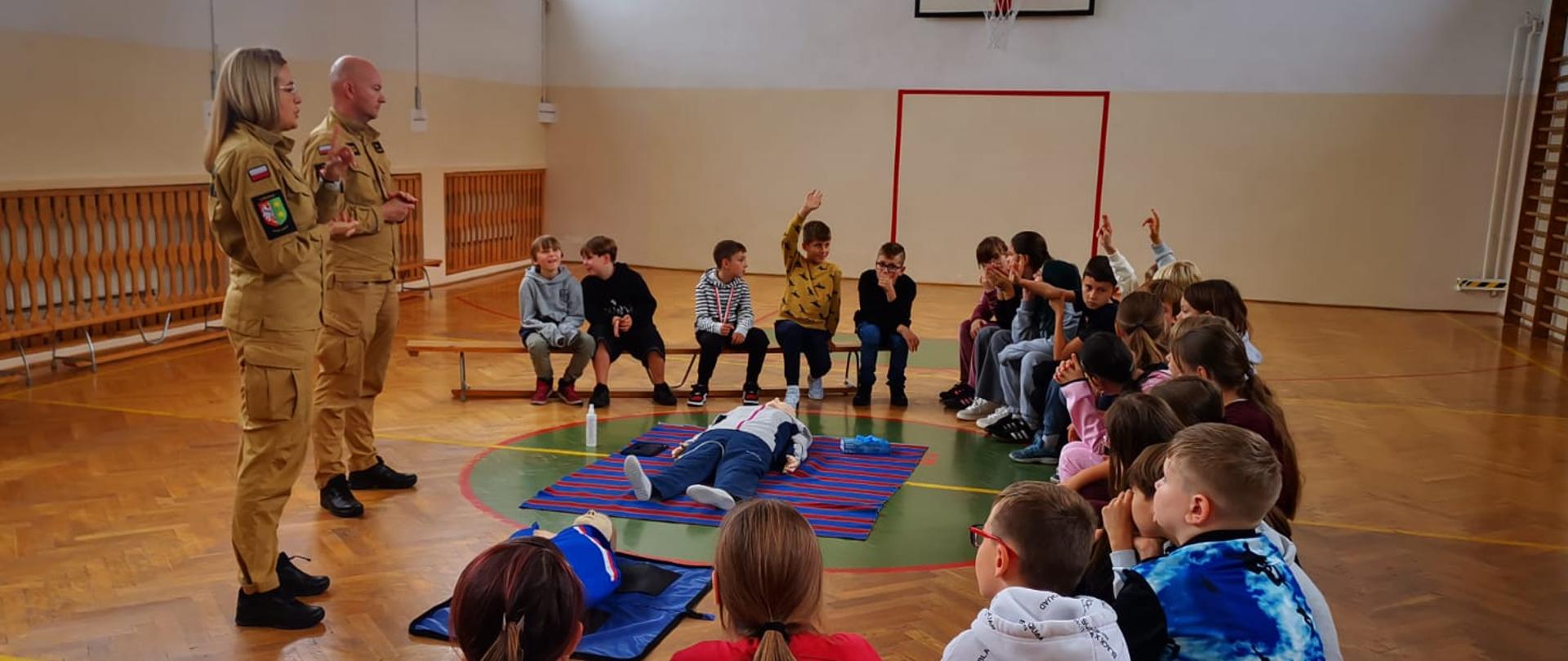 Strażacy prowadza zajęcia edukacyjne z prewencji społecznej na sali gimnastycznej. 