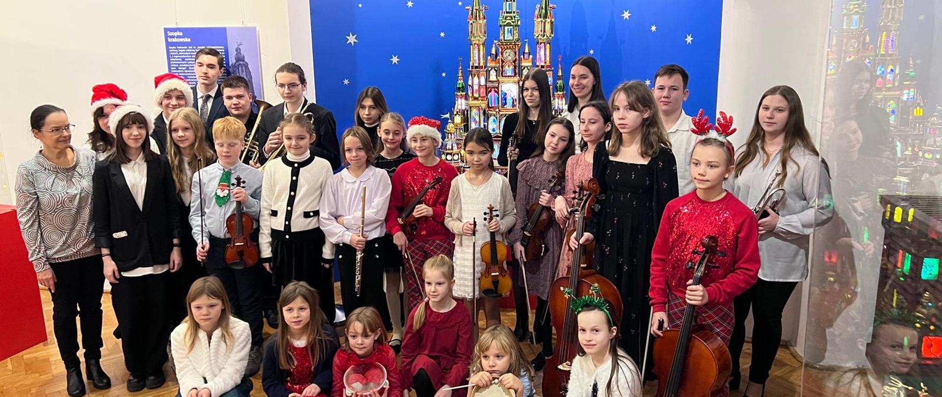Zdjęcie kolorowa. Grupa uczniów z instrumentami stojących na tle szopki noworocznej.