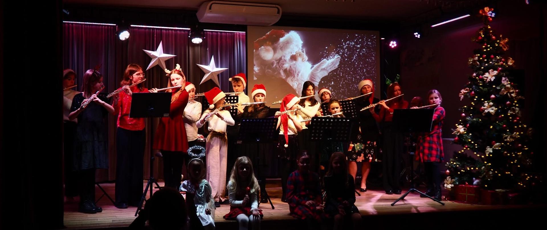 Uczniowie klas fletu występujący na scenie podczas świątecznego koncertu, z akompaniamentem fortepianu, na tle dekoracji bożonarodzeniowych i choinki.
