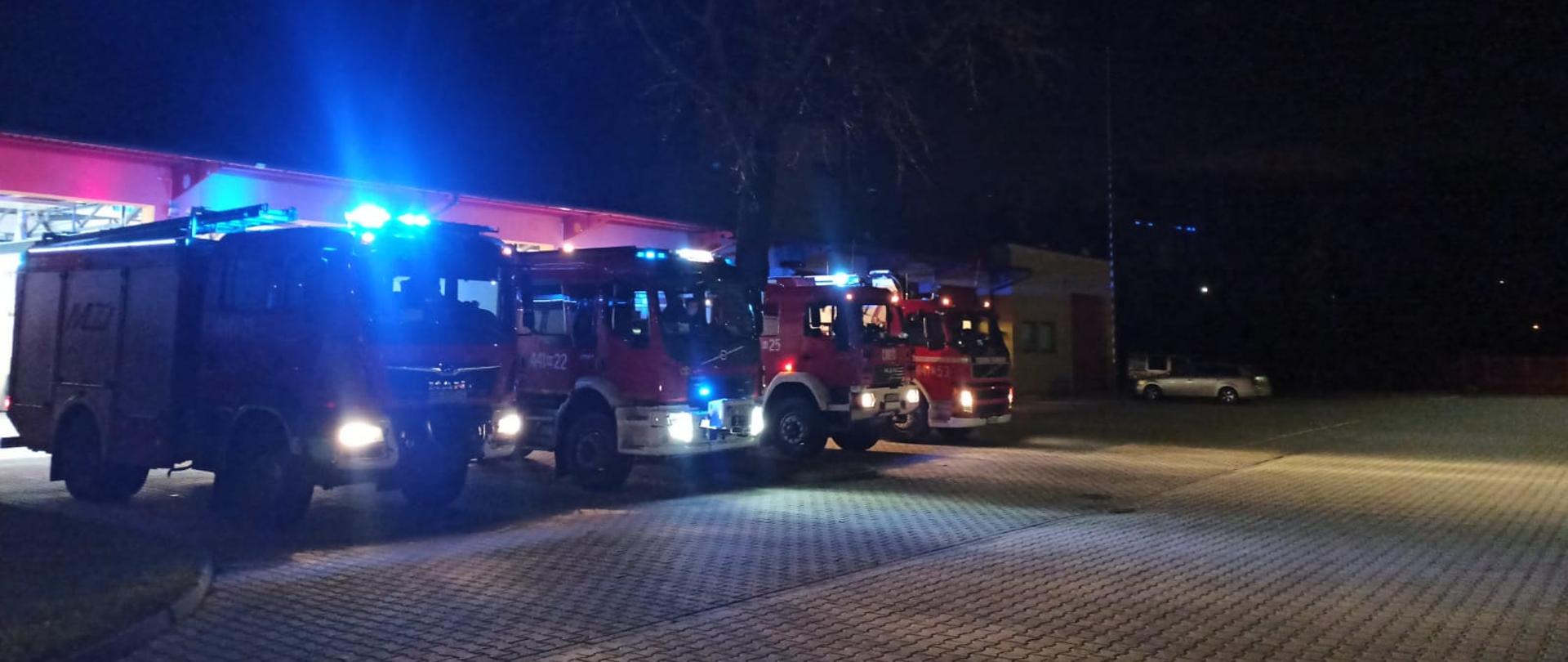 Przed garażami stoja na włączonych sygnałach świetlnych i dźwiękowych samochody pożarnicze w celu oddania hołdu dla tragicznie zmarłych druhów 