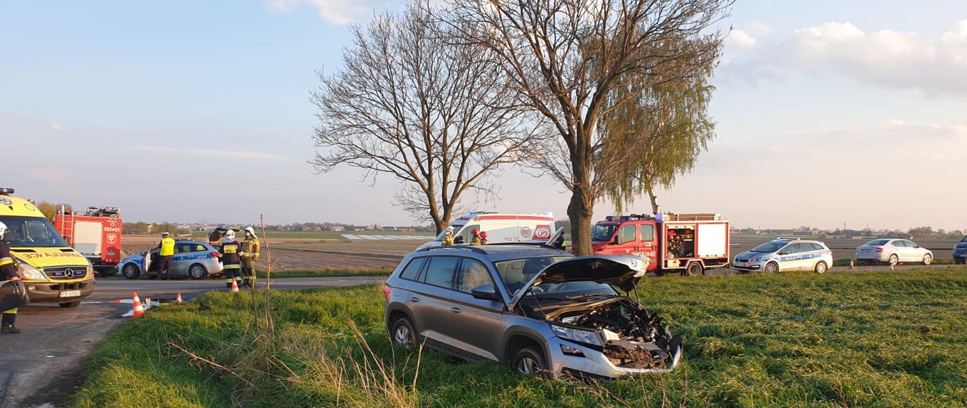 zdjęcie zrobione w czasie wypadku komunikacyjnego. Na jezdni ustawione są pojazdy służb ratunkowych – Straży Pożarnej, Policji i Pogotowia ratunkowego. na polu jest samochód osobowy biorący udział w zderzeniu w wyniku, którego ma uszkodzony przód pojazdu i podniesioną maskę. 