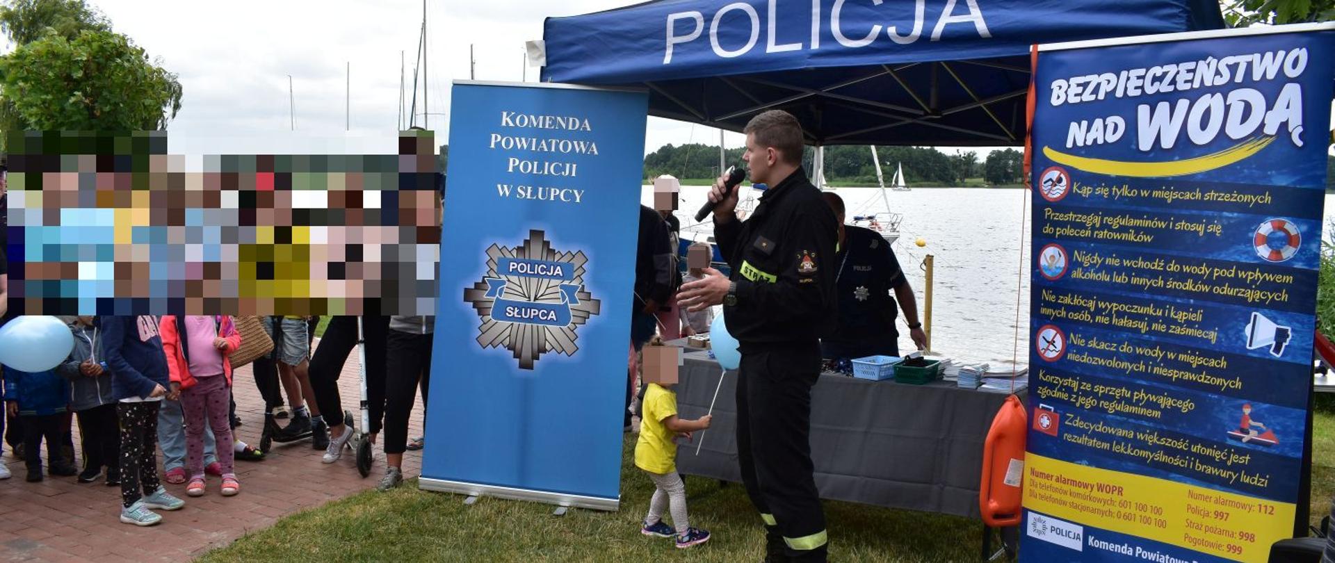 Na zdjęciu strażak z mikrofonem na tle niebieskiego namiotu z napisem policja z boku baner Policji oraz drugi dotyczący akcji prewencyjnej z napisem „bezpieczeństwo nad wodą”. Przed namiotem grupka ludzi wsłuchująca się w prelekcje strażaka. Przy straszaku mała dziewczynka w żółtej koszulce. Przed ludźmi leży na trawie żółta deska ortopedyczna w tle tafla jeziora.