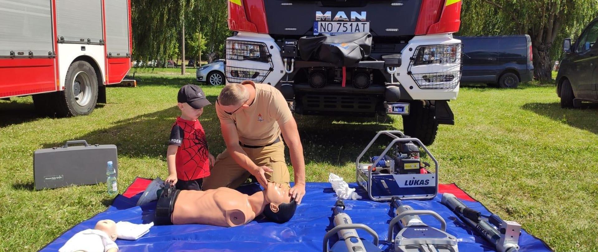 Na trawie rozłożona niebieska mata. Na macie sprzęt strażacki. Obok strażak pokazuje dziecku pierwsza pomoc na manekinie. Za nimi samochód strażacki.