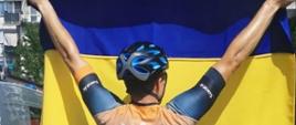 Kolarz ubrany w koszulkę z godłem Ukrainy stoi trzymając flagę Ukrainy. 